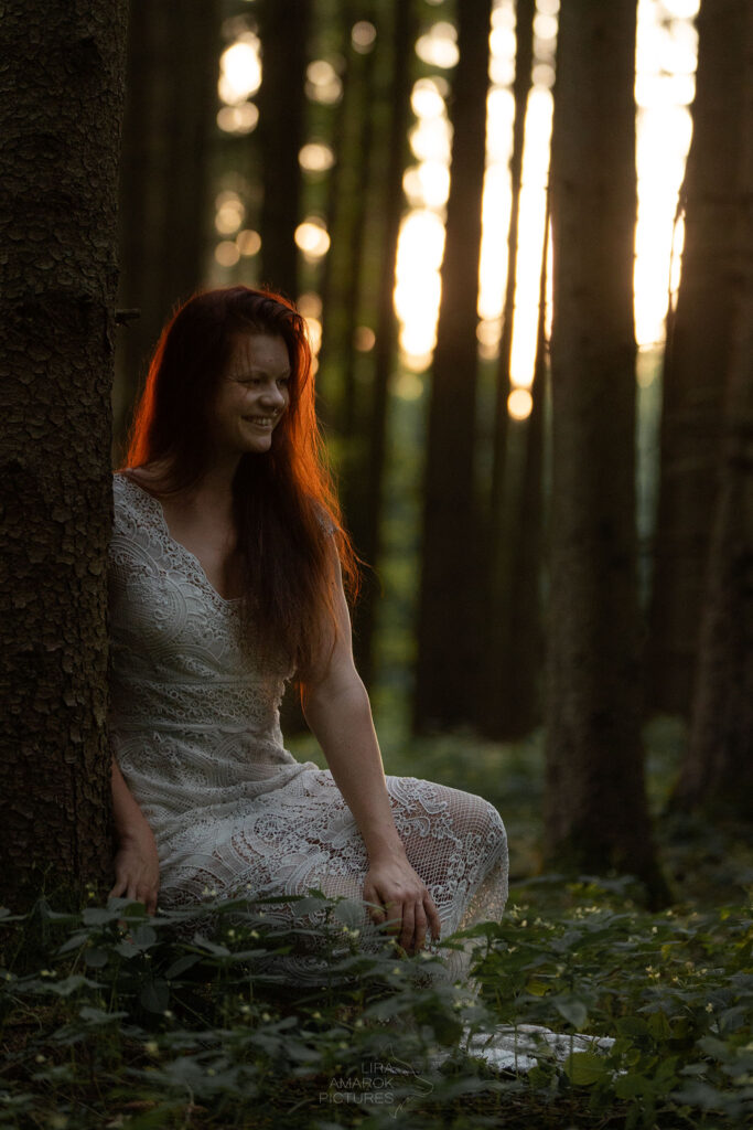 Frau in weißem Kleid sitzt auf Waldboden im Sonnenuntergang
